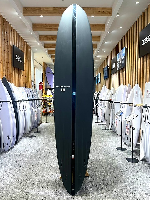 Tall dark blue surfboard standing upright in a surf shop aisle, with white boards on racks along both walls and wooden paneling visible.