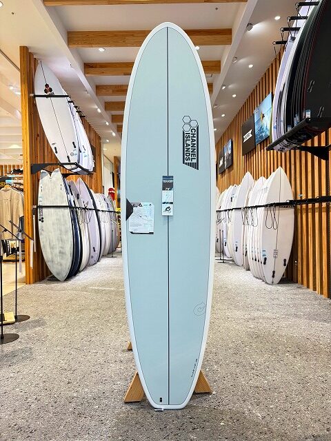 a-image0 - SHOP NEWS | ムラサキスポーツオンラインストア 通販 Light blue longboard surfboard on a wooden stand in a surf shop, with other boards on racks along the walls.