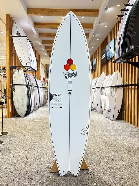 a-image0_1 - SHOP NEWS | ムラサキスポーツオンラインストア 通販 White surfboard on a display stand in a surf shop, centerboard features a red/yellow hex logo and branding; rows of other boards line the walls on both sides.