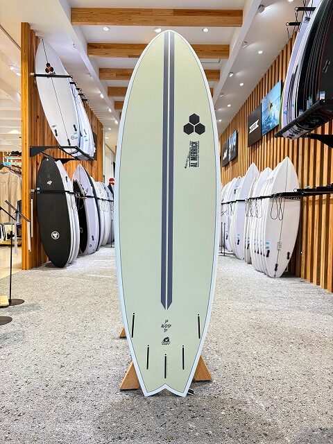 a-image1 - SHOP NEWS | ムラサキスポーツオンラインストア 通販 Pale green longboard surfboard with a dark stripe and hexagonal logo, standing on a wooden rack inside a surf shop with other boards on display.