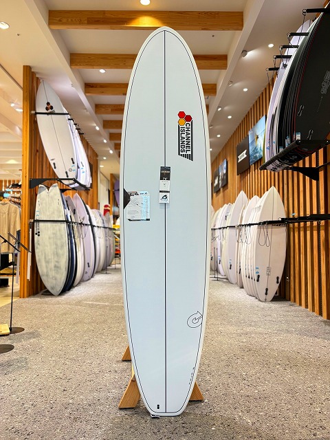 a-image2 - SHOP NEWS | ムラサキスポーツオンラインストア 通販 Light blue surfboard standing upright on a wooden stand in a surf shop, with other boards along the walls.