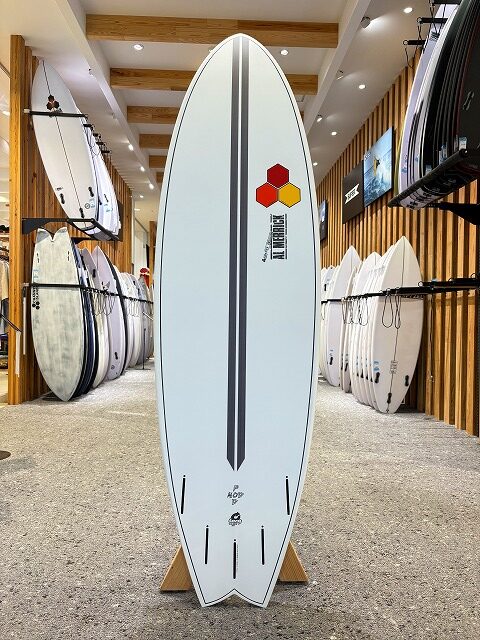 a-image3 - SHOP NEWS | ムラサキスポーツオンラインストア 通販 White surfboard on a stand with a dark vertical stripe and hexagon logo, displayed in a surf shop.