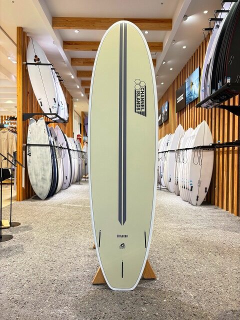 a-image3 - SHOP NEWS | ムラサキスポーツオンラインストア 通販 Pale yellow surfboard with a dark vertical stripe on a stand in a surf shop showroom, surrounded by other boards on racks.