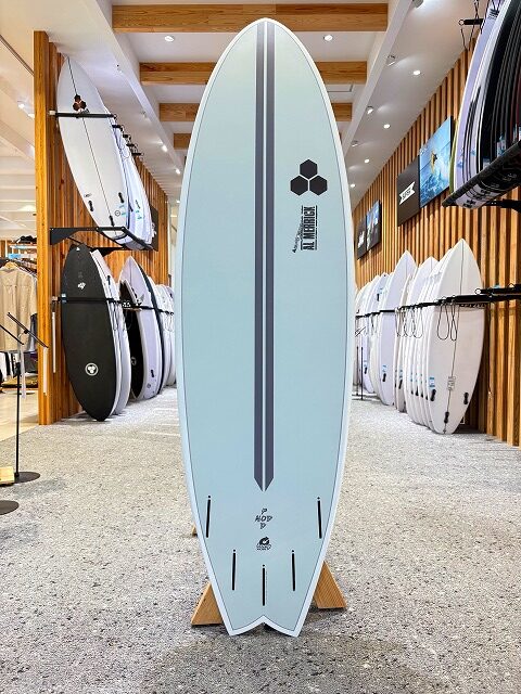 a-image4 - SHOP NEWS | ムラサキスポーツオンラインストア 通販 Light blue surfboard on a display stand inside a shop, surrounded by other boards and racks.