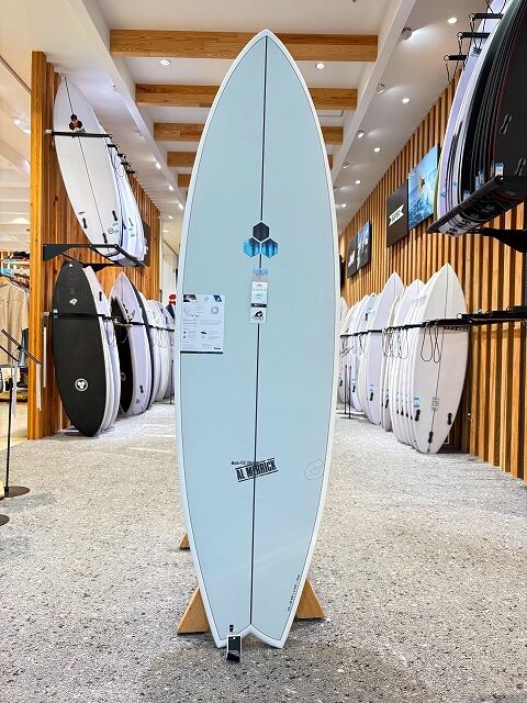a-image5 - SHOP NEWS | ムラサキスポーツオンラインストア 通販 Light blue surfboard on a display stand in a surf shop showroom, with rows of surfboards on racks along wooden walls.