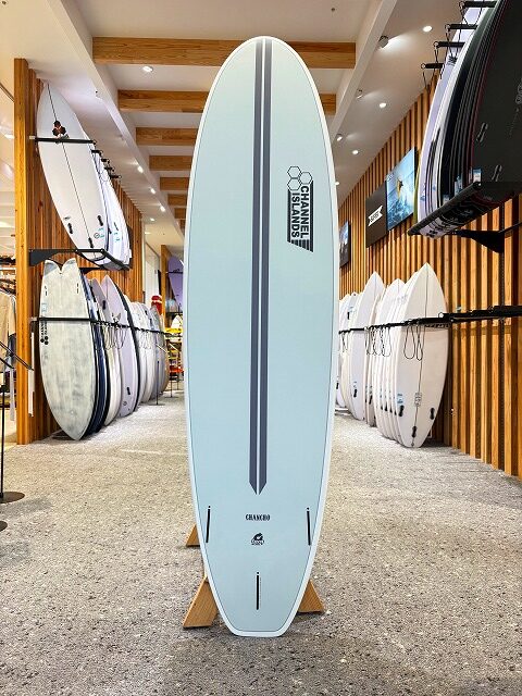 a-image5 - SHOP NEWS | ムラサキスポーツオンラインストア 通販 Light blue surfboard with a vertical dark stripe on a wooden stand inside a surf shop, logo visible, surrounded by other boards on racks.