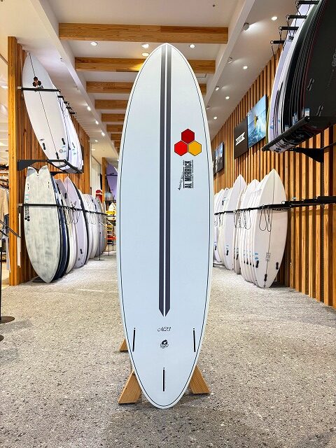 a-image6 - SHOP NEWS | ムラサキスポーツオンラインストア 通販 White surfboard on a wooden stand in a surf shop, featuring a black center stripe and logo near the top, with other boards on racks around it.