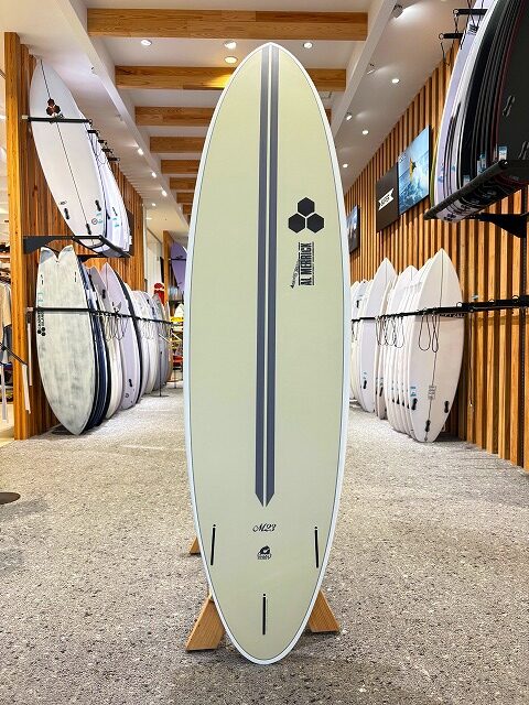 a-image8 - SHOP NEWS | ムラサキスポーツオンラインストア 通販 Cream surfboard with dark dual stripes and a central line, upright on a wooden stand in a surf shop, with racks of boards along the walls.