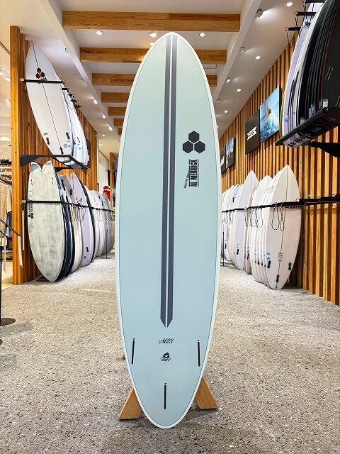 a-image9 - SHOP NEWS | ムラサキスポーツオンラインストア 通販 Light blue surfboard on a wooden stand inside a surf shop, with racks of boards lining both sides in the background.