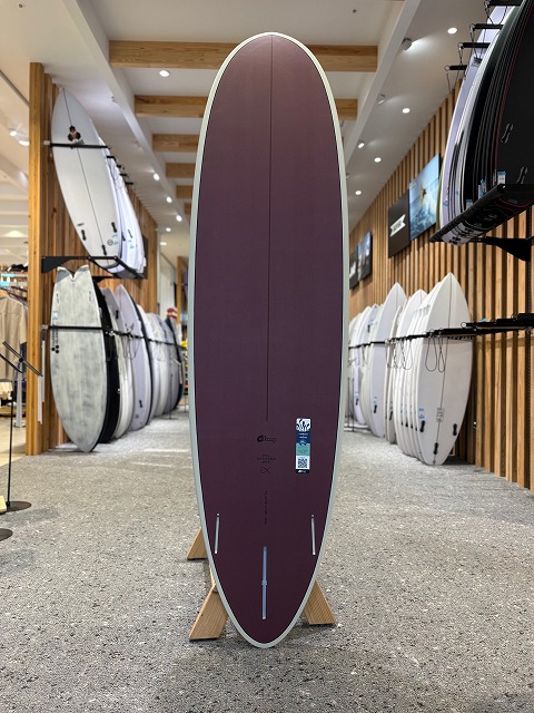 b-image1 - SHOP NEWS | ムラサキスポーツオンラインストア 通販 Purple surfboard standing upright on a wooden display stand inside a surf shop, with other boards on racks along the walls.