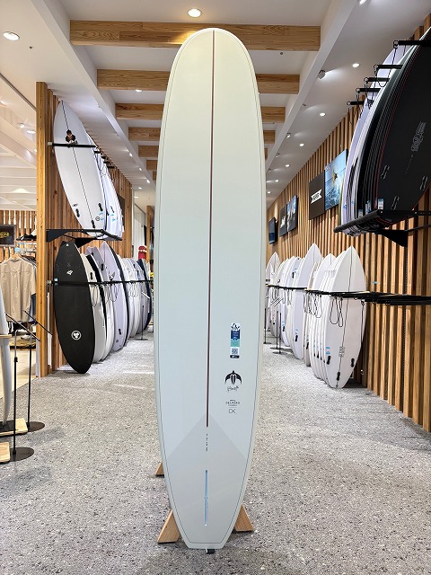 b-image3 - SHOP NEWS | ムラサキスポーツオンラインストア 通販 Tall white surfboard with a red stringer standing upright on a wooden rack in a surf shop, surrounded by other boards.
