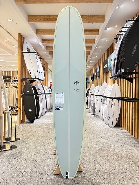 b-image4 - SHOP NEWS | ムラサキスポーツオンラインストア 通販 Tall pale green surfboard on a stand in a surf shop showroom, surrounded by other boards on racks.