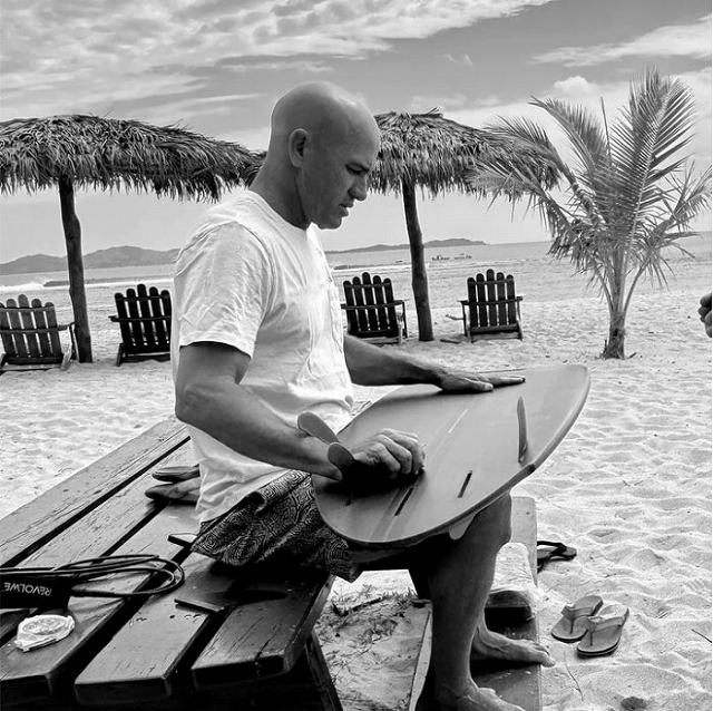 s-スクリーンショット 2026-04-27 192047 - SHOP NEWS | ムラサキスポーツオンラインストア 通販 Bald man in a white t-shirt sits on a wooden bench on a beach, shaping a surfboard with a plane.