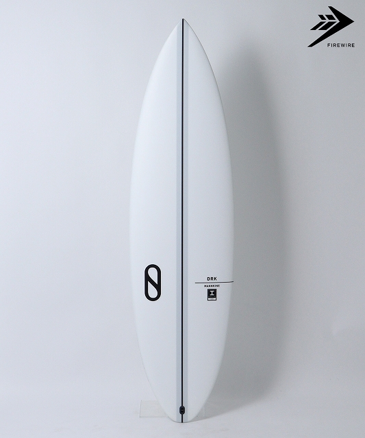 s-DRK - SHOP NEWS | ムラサキスポーツオンラインストア 通販 White surfboard standing upright against a pale wall, with a black logo on its left and a Firewire mark in the top-right corner.