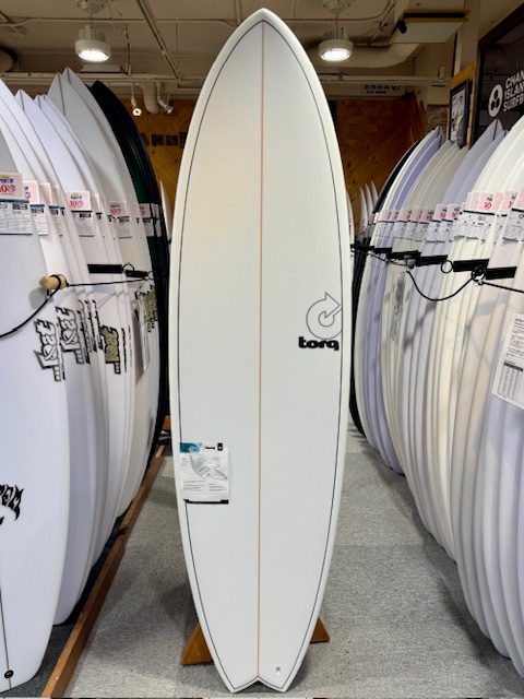White surfboard displayed upright between rows of other boards in a shop, featuring an orange centerline and a Torq logo on the right side.