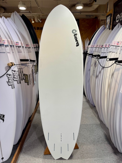 White surfboard on display in a shop, with a Globe logo on the upper right, surrounded by other surfboards on racks.