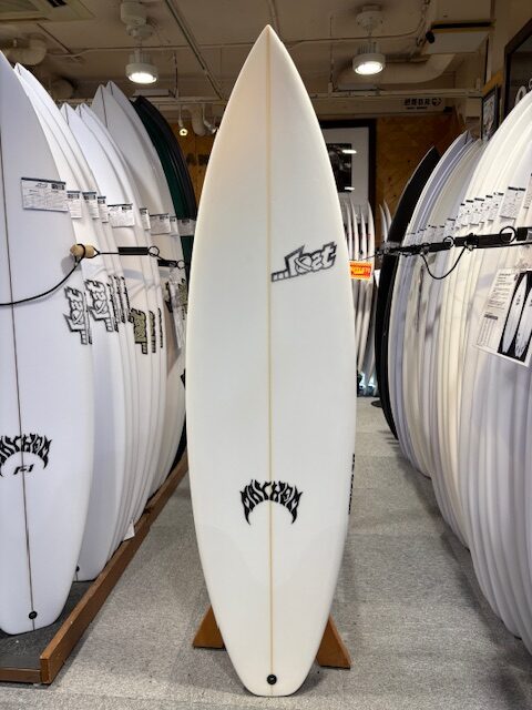 White surfboard on a stand in a surf shop, surrounded by racks of other boards.