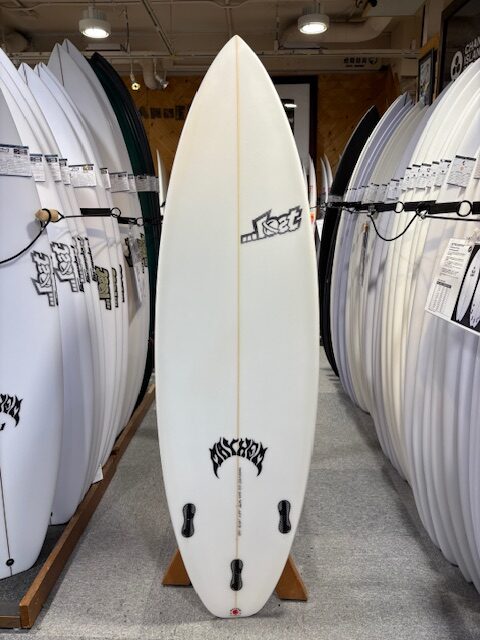 White surfboard standing upright on a rack in a shop, with twin fins and logos near the nose and tail.