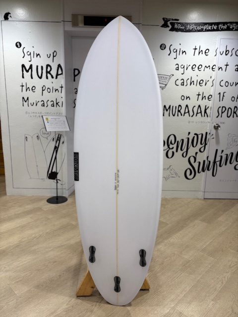 White surfboard on a wooden rack indoors, fin boxes visible at the tail, with graffiti-style surf shop walls in the background.