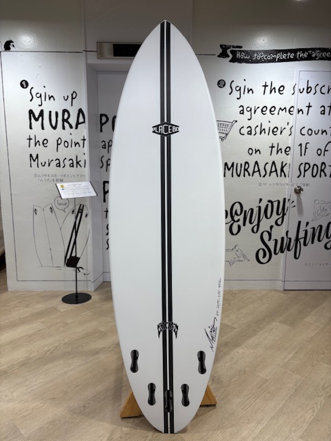 IMG_7874 - SHOP NEWS | ムラサキスポーツオンラインストア 通販 White surfboard with a black center stripe and logo, upright on a stand, showing four fin boxes near the bottom in a shop with graffiti-style walls.