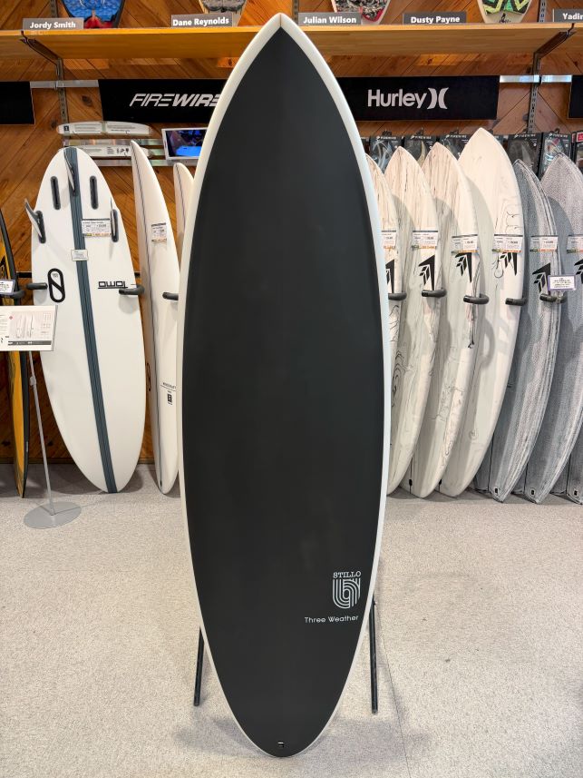 Black surfboard with white rails on a display stand in a surf shop, surrounded by white and gray boards in the background.
