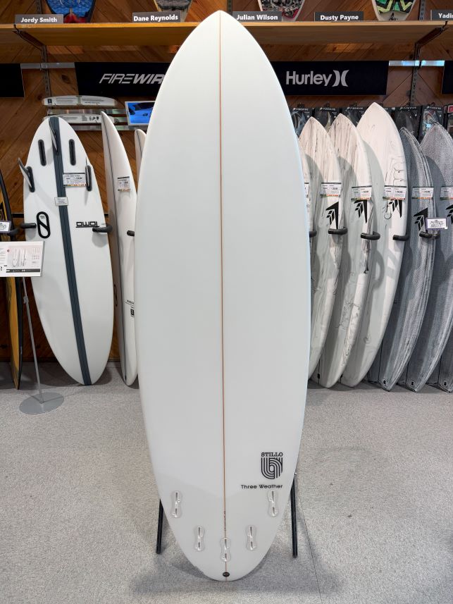Large white surfboard on a display stand in a shop, surrounded by other boards and brand banners, including Hurley, in the background.