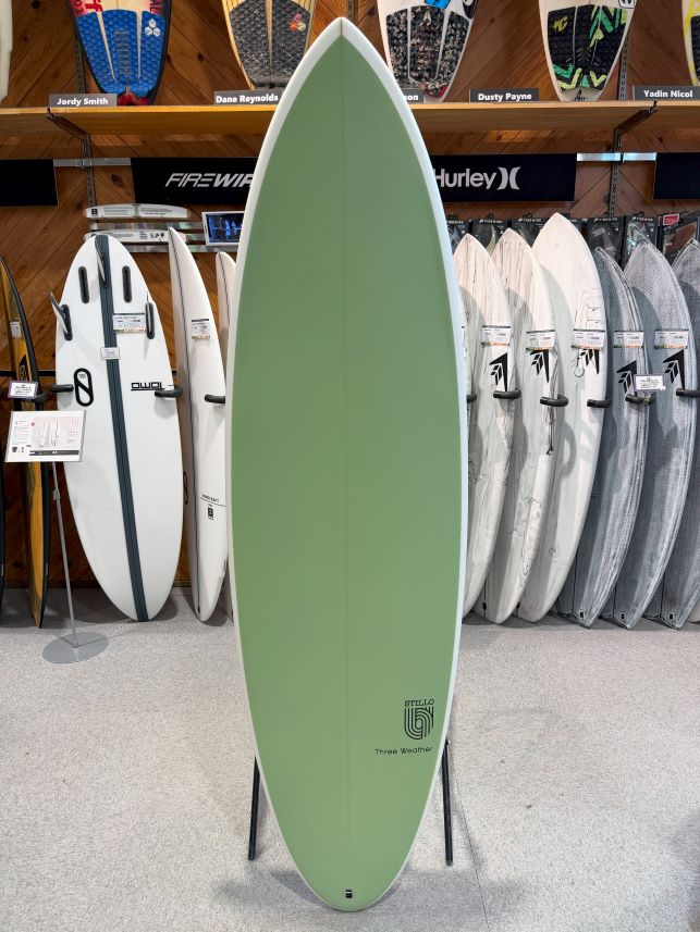 Green surfboard on a display stand in a surf shop, with white and gray boards on racks behind it.