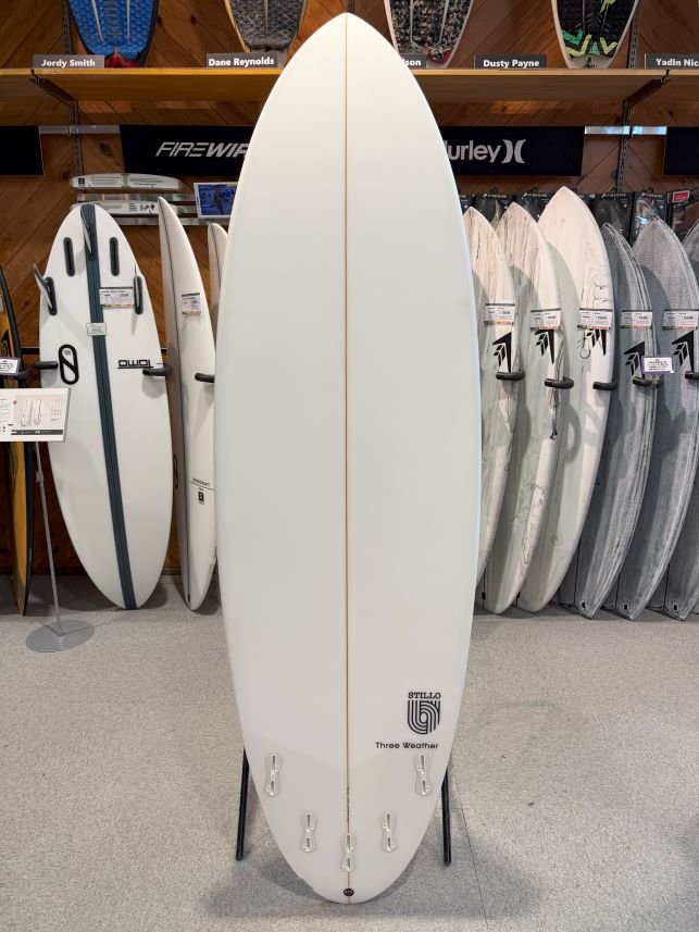 White surfboard on a display rack in a surf shop, standing upright with a light brown stringer and Three Weather logo near the tail, surrounded by other boards on shelves.