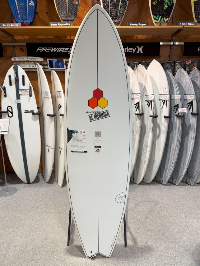 White surfboard on a display stand in a shop, center stage with hexagon logo and 'AL MARRICK' text, surrounded by otherboards on racks.