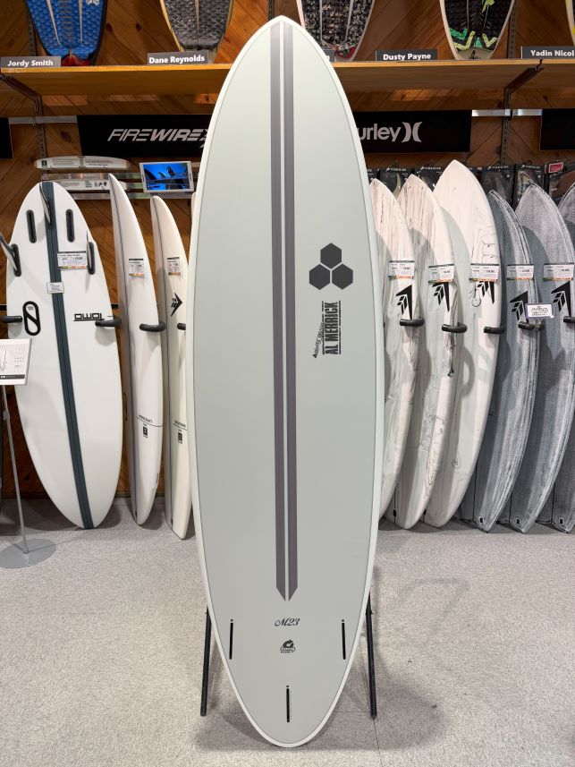 Tall white surfboard on a display rack in a shop, with gray center stripe and hex logo, surrounded by other boards on racks behind it.