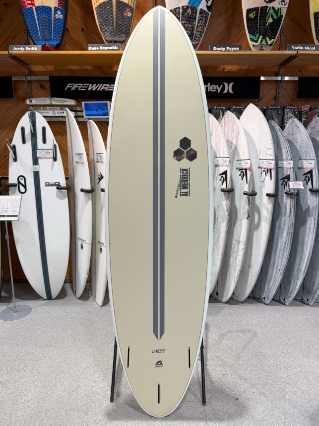 Beige longboard on a display stand inside a surf shop, with other boards on racks in the background.