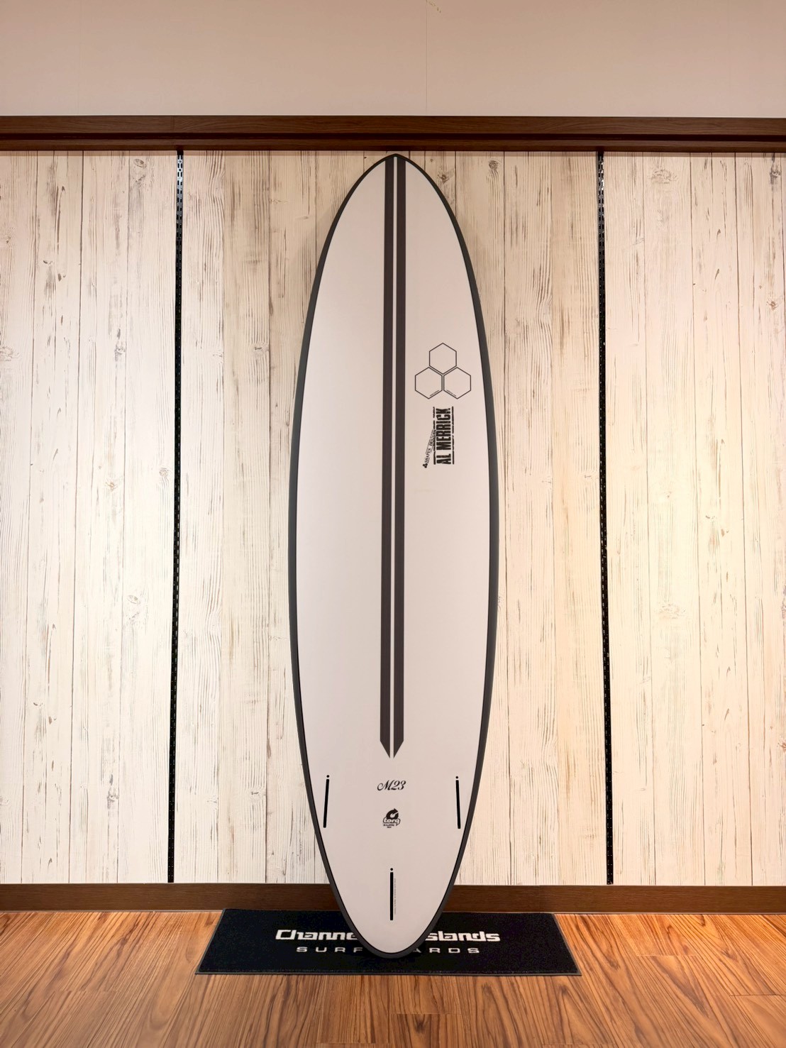 White surfboard with a black central stripe, mounted vertically on a white wood-paneled wall.