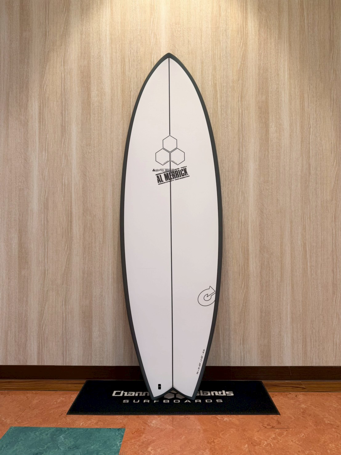 White surfboard with black border standing upright against a wood-paneled wall on a display mat.