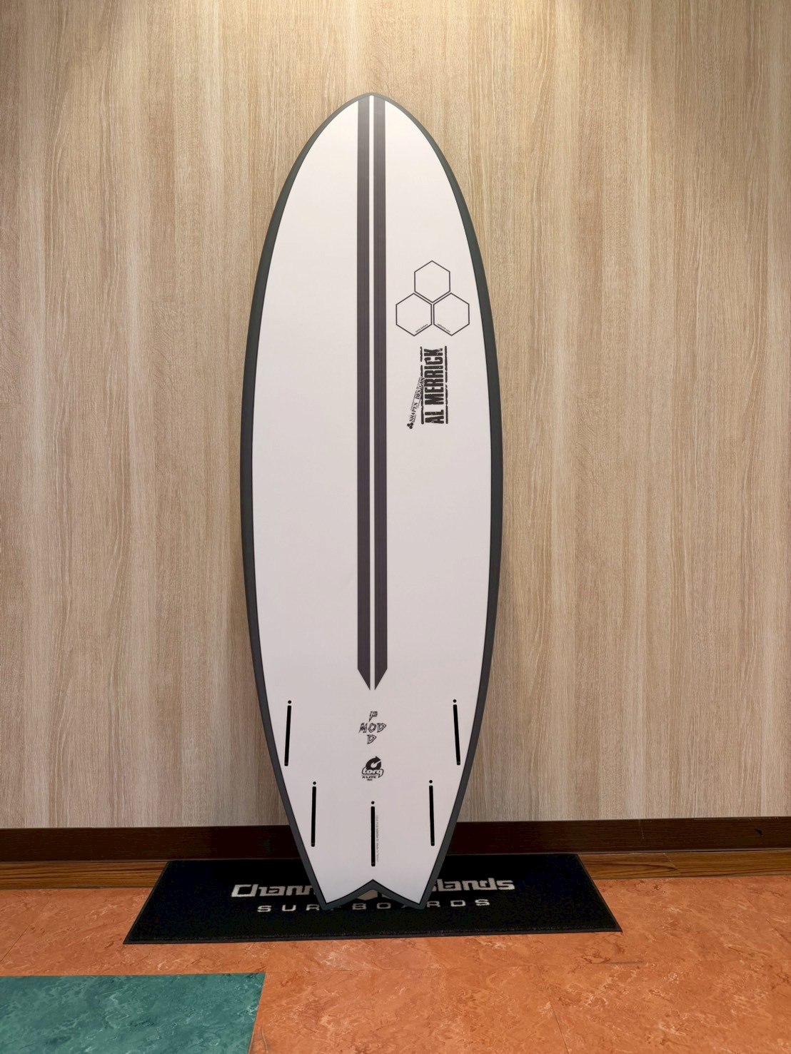 White surfboard with double black center stripes and hexagon motif, leaning against a light wood wall on a black-mat display.