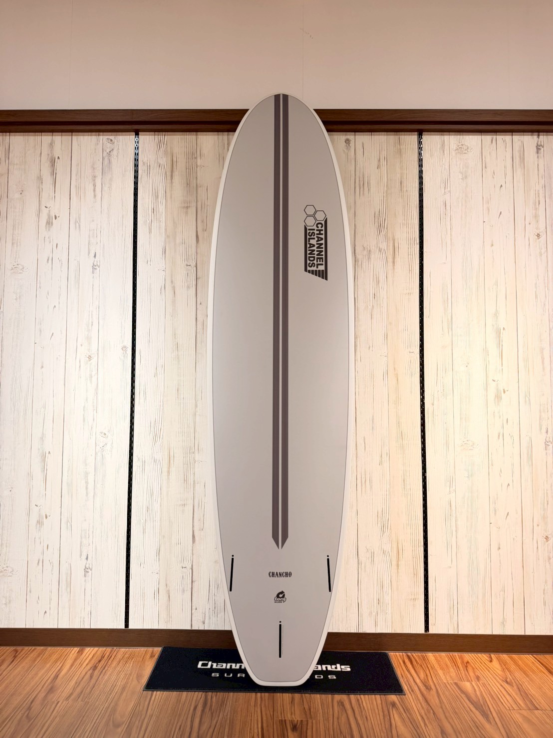 White surfboard standing upright indoors on a display mat, with a vertical dark stripe and Channel Islands logo near the top.