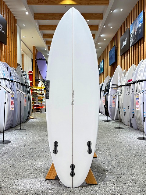 White surfboard on a display rack in a surf shop, centered with other boards lining the aisle behind it.