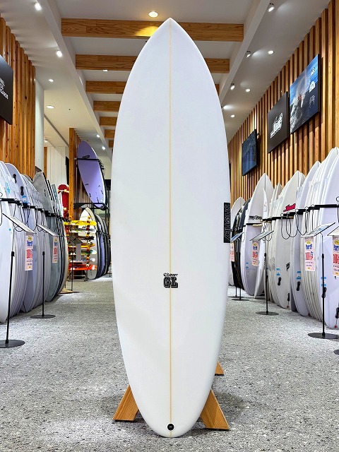 White longboard standing upright on a wooden rack inside a surf shop, with many other boards lining the walls.