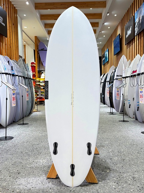 White surfboard standing upright on a wooden display stand in a surf shop, flanked by rows of boards on racks.