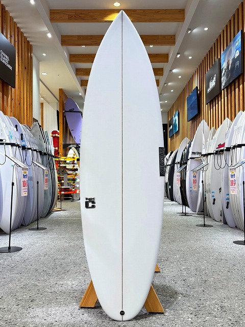 White surfboard standing upright on a wooden rack in a surf shop, with rows of boards lining the walls on both sides.