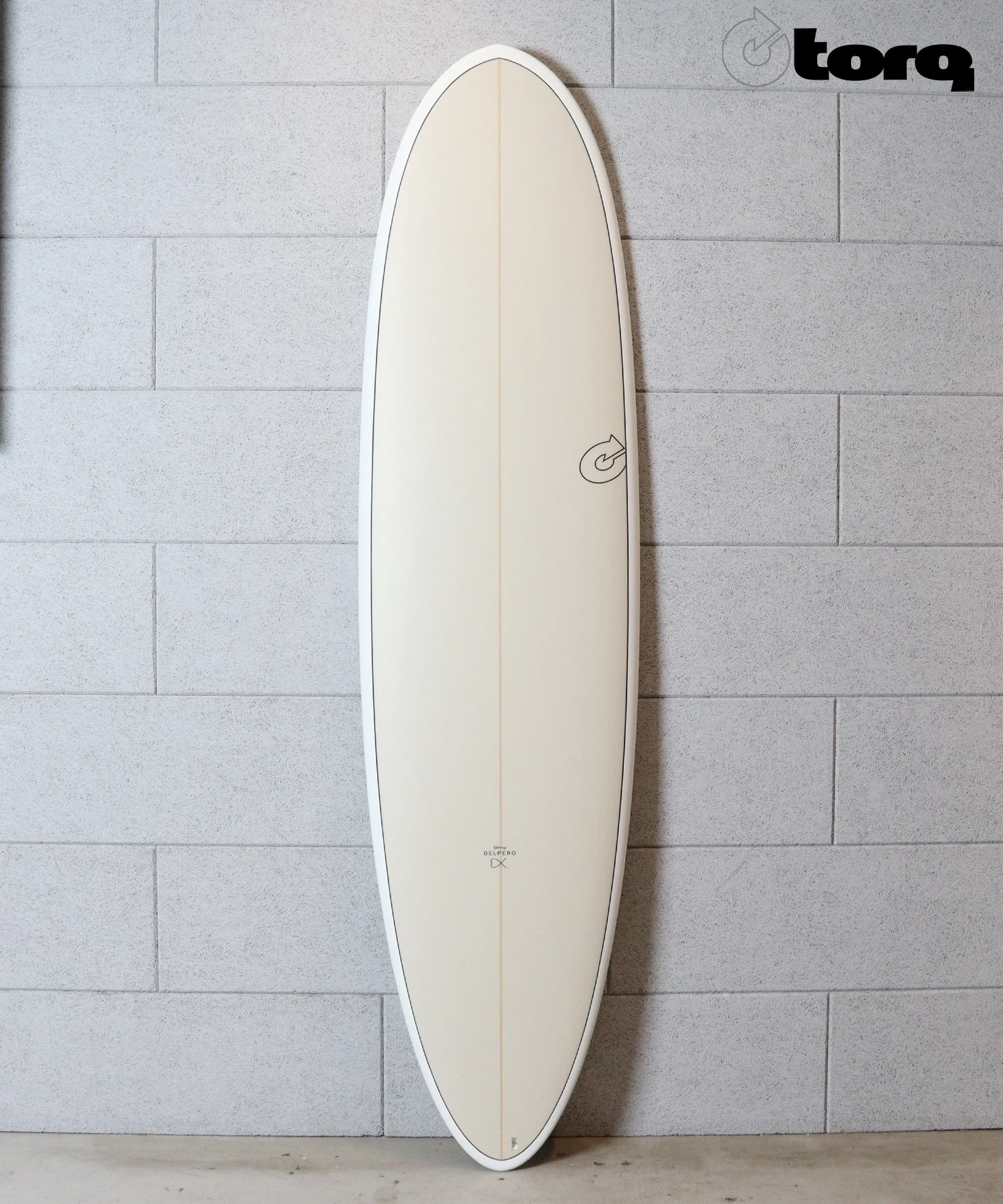 Beige longboard surfboard standing upright indoors against a gray tiled wall, with a small logo near the bottom.