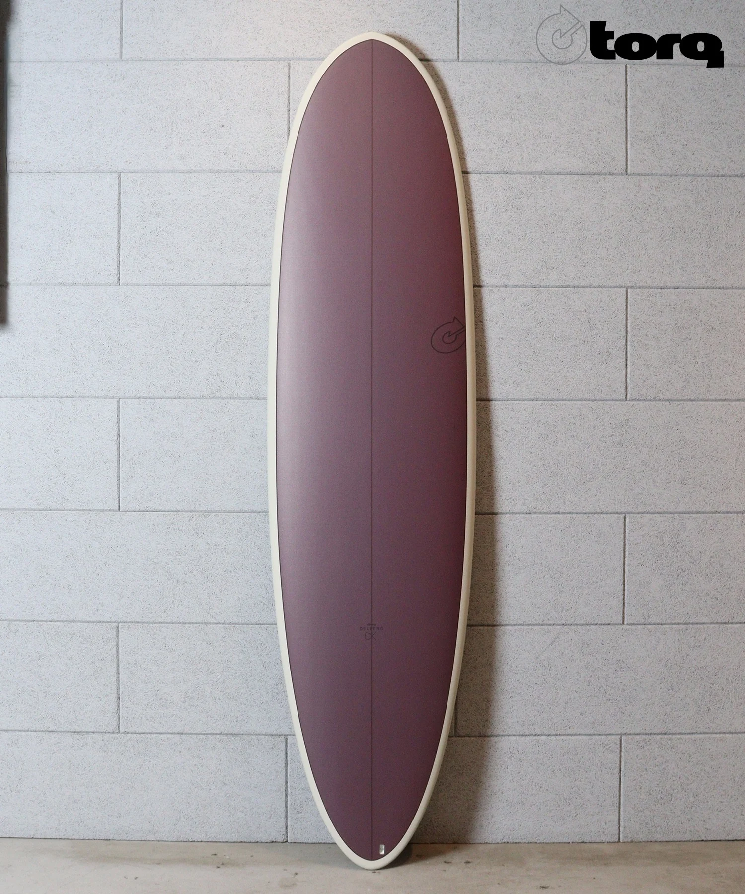 Purple surfboard with a cream edge standing upright against a gray cinder block wall, Torq logo in the corner.