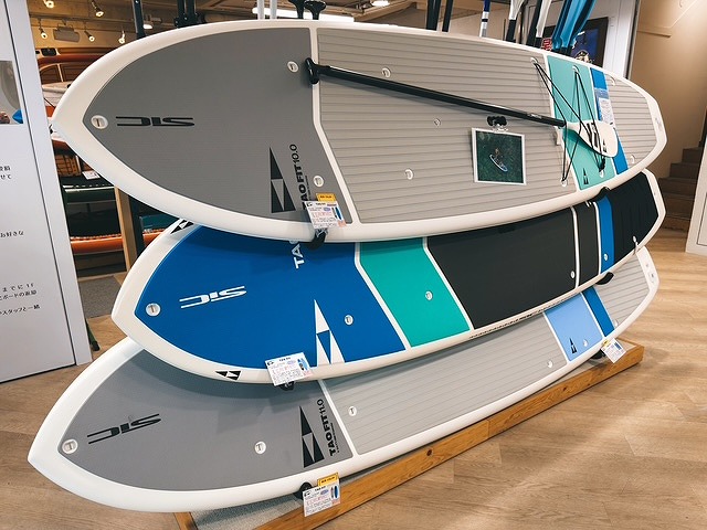 s-81B8A99D-8EAA-449E-9037-EF7AFC9E9DDF - SHOP NEWS | ムラサキスポーツオンラインストア 通販 Stack of stand-up paddleboards on a showroom display rack, gray, blue, teal, and black boards with logos visible.