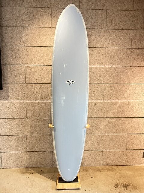 Light blue surfboard standing upright on a wooden rack indoors against a beige brick wall.