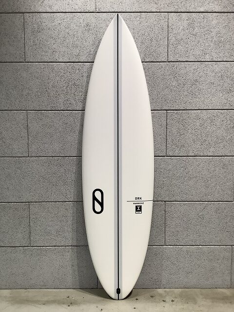 s-IMG_6434 - SHOP NEWS | ムラサキスポーツオンラインストア 通販 White surfboard standing upright against a gray concrete block wall, showing a dark center stringer and a black logo toward the tail.