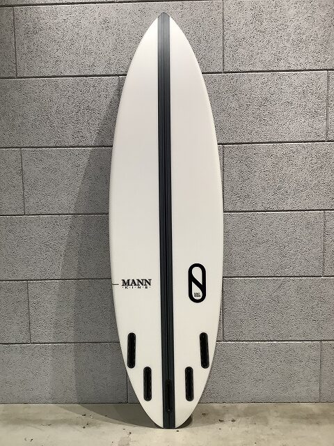 s-IMG_6435 - SHOP NEWS | ムラサキスポーツオンラインストア 通販 White surfboard with a black center stringer and four fin boxes, standing against a gray cinder block wall; features Mann branding and a black oval logo on the right.