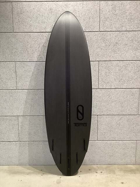 s-IMG_6437 - SHOP NEWS | ムラサキスポーツオンラインストア 通販 Dark gray surfboard standing upright against a light gray brick wall, with a center groove and four fin boxes at the bottom, featuring a 'TOMO' logo.