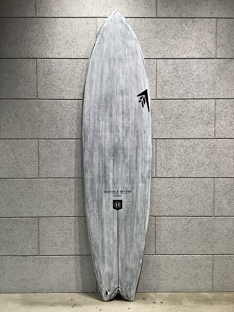s-IMG_6438 - SHOP NEWS | ムラサキスポーツオンラインストア 通販 Grey surfboard with faint wood grain, standing upright against a light gray tiled wall; visible logos near the center and tail area.