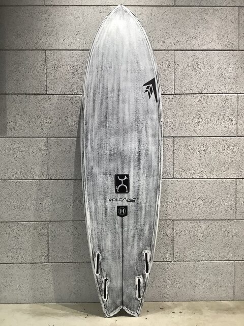 s-IMG_6439 - SHOP NEWS | ムラサキスポーツオンラインストア 通販 Gray surfboard standing upright against a tiled wall, featuring brand logos along its deck.
