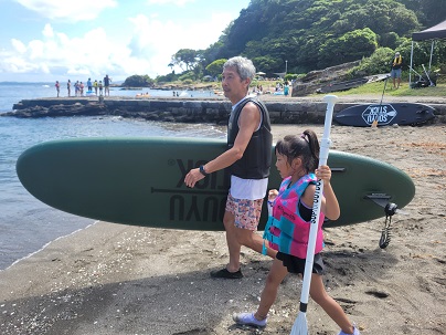 Man and a girl carry a green surfboard toward the water on a sandy beach with rocky shoreline and calm blue sea behind them.
