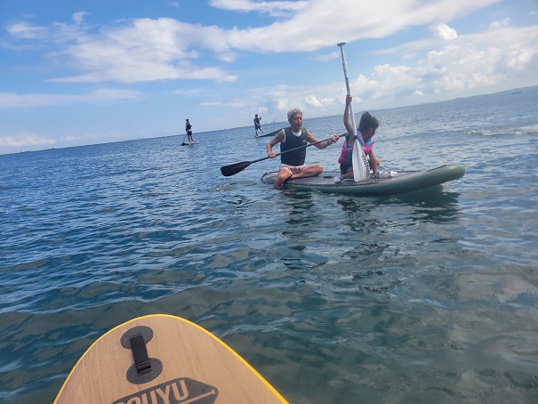 View from a paddleboard: two people on a stand-up board in blue water, one raising a paddle while others paddle nearby.